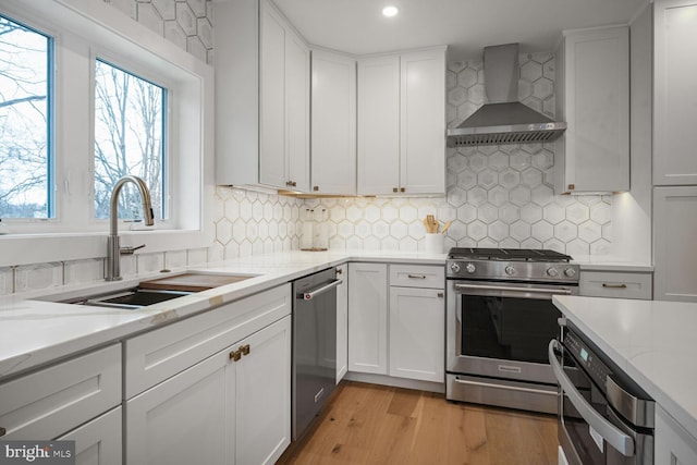 kitchen with stainless steel appliances, white cabinets, a sink, wall chimney range hood, and light stone countertops