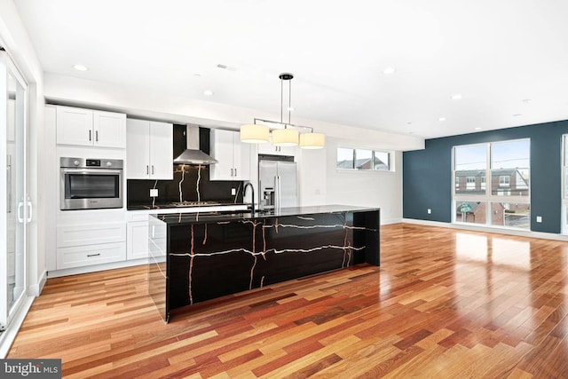 kitchen with decorative light fixtures, an island with sink, white cabinetry, stainless steel appliances, and wall chimney range hood