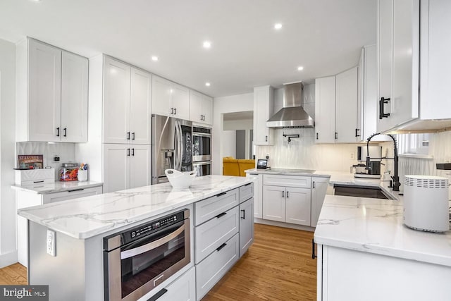 kitchen featuring wall chimney range hood, sink, appliances with stainless steel finishes, white cabinetry, and a center island