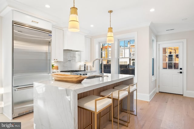 kitchen featuring built in fridge, hanging light fixtures, white cabinetry, and a center island with sink