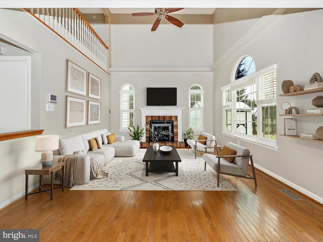 living room featuring ceiling fan, a brick fireplace, a towering ceiling, and light hardwood / wood-style floors