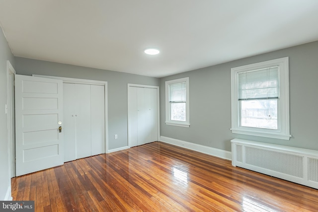 unfurnished bedroom with wood finished floors, radiator heating unit, two closets, and baseboards