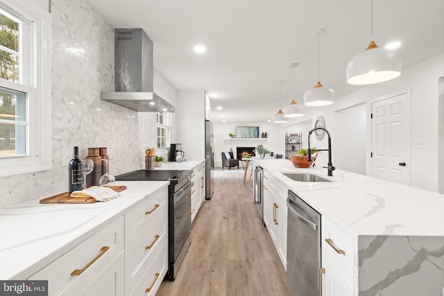 kitchen featuring appliances with stainless steel finishes, a sink, a lit fireplace, wall chimney range hood, and backsplash