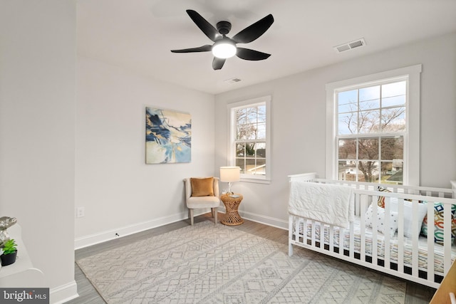 bedroom with wood finished floors, a ceiling fan, visible vents, baseboards, and a crib