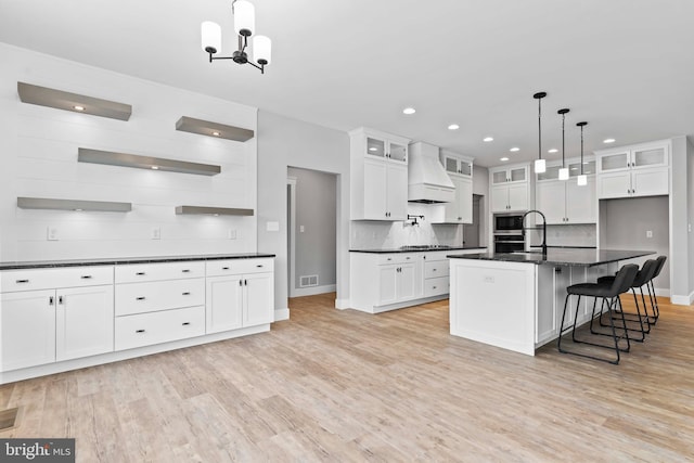 kitchen with pendant lighting, white cabinetry, light hardwood / wood-style floors, an island with sink, and custom exhaust hood