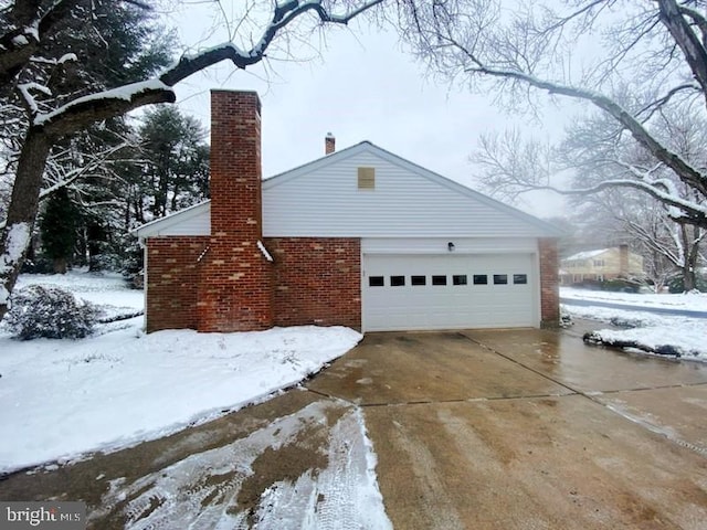 view of snowy exterior featuring driveway, an attached garage, a chimney, and brick siding