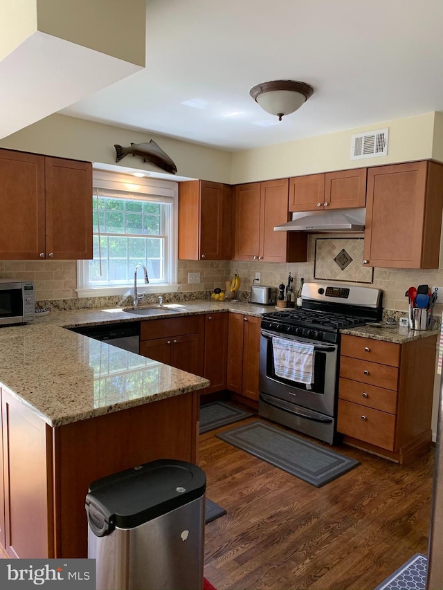 kitchen with appliances with stainless steel finishes, a sink, under cabinet range hood, brown cabinetry, and dark wood-type flooring