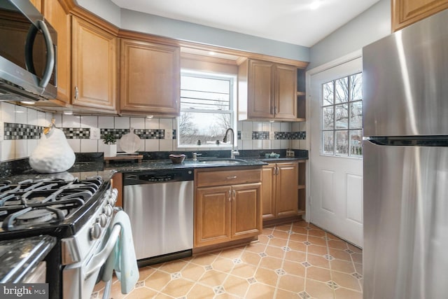 kitchen with appliances with stainless steel finishes, brown cabinets, dark stone countertops, a sink, and backsplash