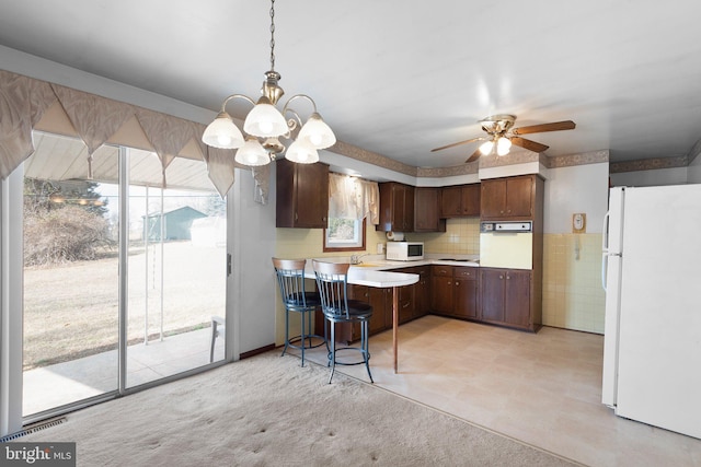 kitchen with ceiling fan with notable chandelier, pendant lighting, kitchen peninsula, dark brown cabinets, and white appliances