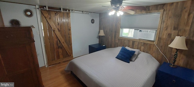 bedroom with wooden walls, cooling unit, hardwood / wood-style flooring, ceiling fan, and a barn door