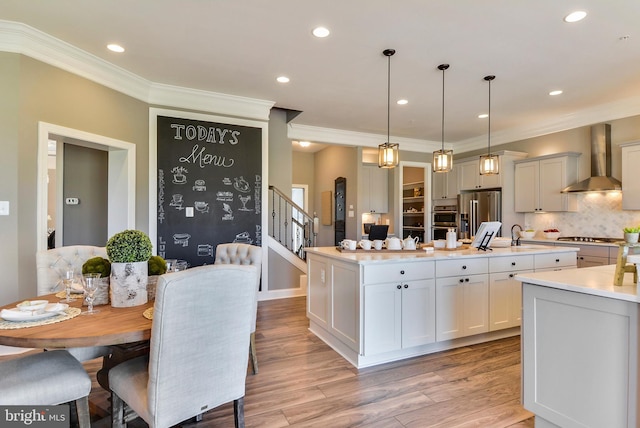 kitchen featuring decorative light fixtures, a center island with sink, appliances with stainless steel finishes, wall chimney range hood, and white cabinets