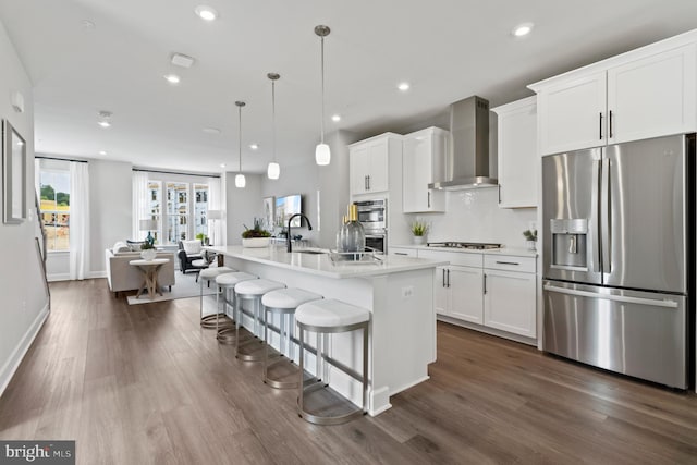 kitchen with wall chimney range hood, pendant lighting, stainless steel appliances, a kitchen island with sink, and white cabinets