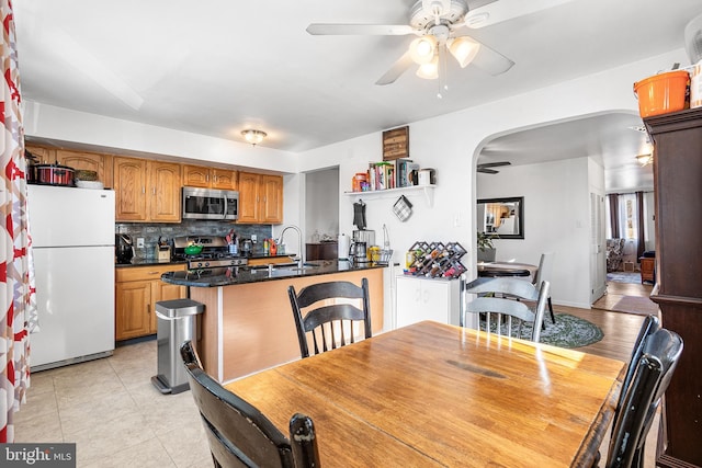 kitchen featuring arched walkways, light tile patterned floors, appliances with stainless steel finishes, brown cabinetry, and ceiling fan