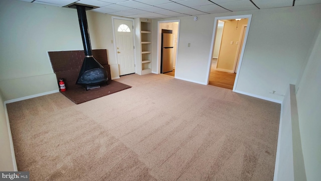 basement with stainless steel refrigerator, a drop ceiling, built in shelves, light colored carpet, and a wood stove