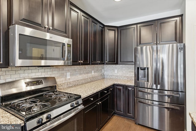 kitchen with appliances with stainless steel finishes, decorative backsplash, dark brown cabinetry, light stone countertops, and light hardwood / wood-style flooring