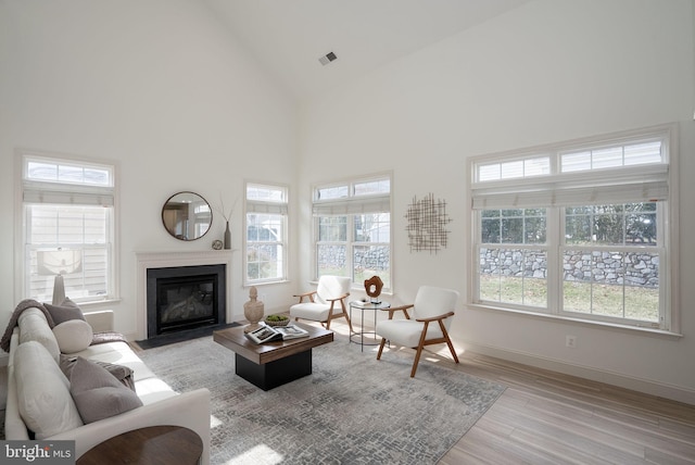 living area featuring visible vents, a fireplace with flush hearth, high vaulted ceiling, light wood-type flooring, and baseboards