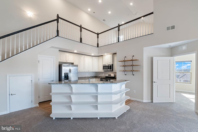 kitchen featuring dark stone countertops, a towering ceiling, stainless steel appliances, white cabinets, and light colored carpet