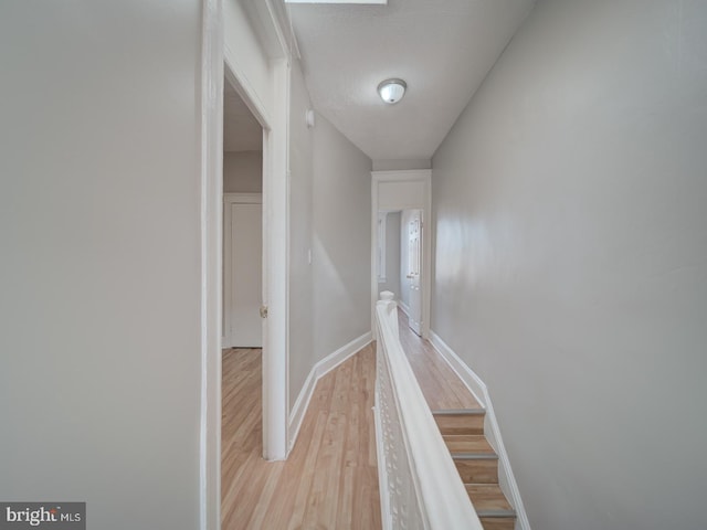 corridor featuring light wood-style flooring, baseboards, a textured ceiling, and an upstairs landing