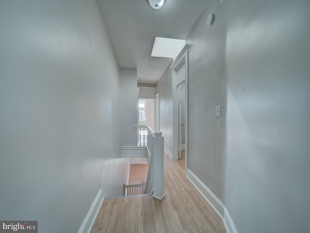 corridor with a textured ceiling, light wood-type flooring, an upstairs landing, and baseboards