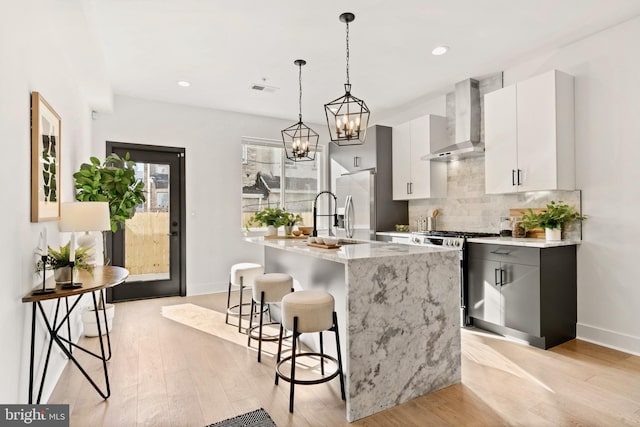 kitchen with pendant lighting, wall chimney range hood, appliances with stainless steel finishes, a kitchen island with sink, and white cabinetry