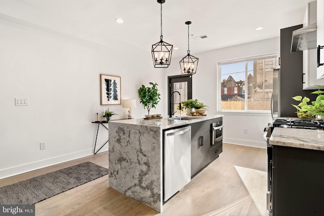 kitchen with sink, appliances with stainless steel finishes, light stone countertops, a center island with sink, and decorative light fixtures