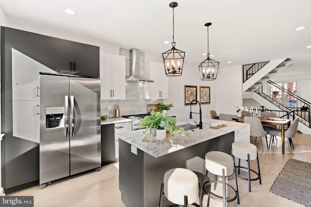 kitchen featuring sink, wall chimney range hood, stainless steel appliances, an island with sink, and decorative light fixtures