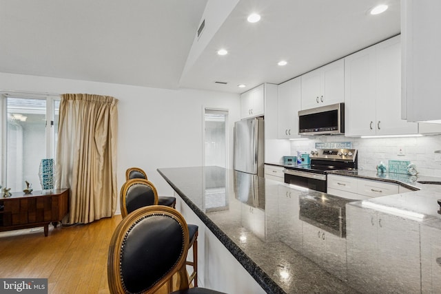 kitchen with white cabinets, appliances with stainless steel finishes, backsplash, light wood finished floors, and dark stone countertops