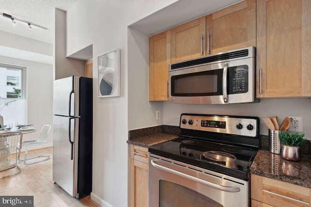 kitchen with rail lighting, appliances with stainless steel finishes, dark stone countertops, light brown cabinets, and light wood-type flooring