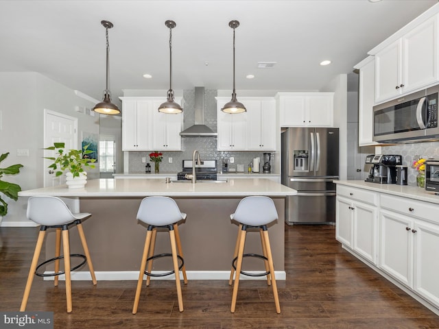 kitchen with appliances with stainless steel finishes, white cabinets, hanging light fixtures, a center island with sink, and wall chimney exhaust hood