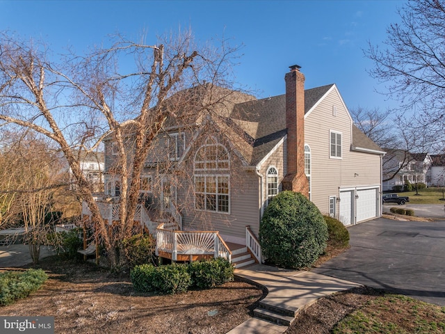 view of front of property featuring a wooden deck, an attached garage, a shingled roof, a chimney, and aphalt driveway