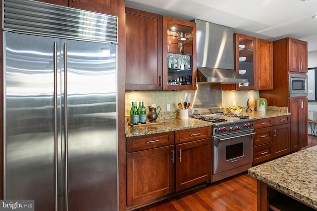 kitchen with dark hardwood / wood-style flooring, decorative backsplash, built in appliances, light stone counters, and wall chimney exhaust hood