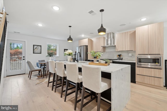 kitchen featuring hanging light fixtures, light brown cabinetry, an island with sink, stainless steel appliances, and wall chimney range hood