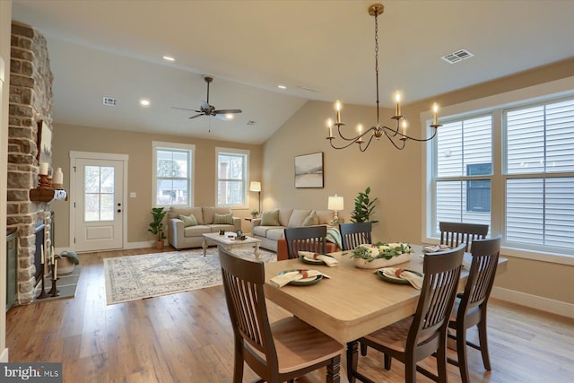 dining area with ceiling fan with notable chandelier, lofted ceiling, a stone fireplace, and light hardwood / wood-style floors