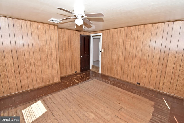 empty room with a ceiling fan, visible vents, dark wood finished floors, and wooden walls