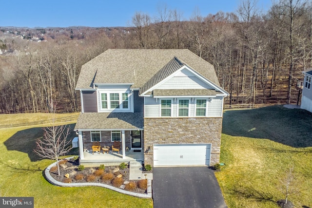 view of front facade featuring driveway, stone siding, a wooded view, and a front yard