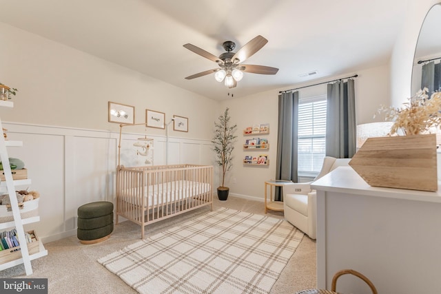 bedroom with a crib, visible vents, a ceiling fan, light colored carpet, and a wainscoted wall