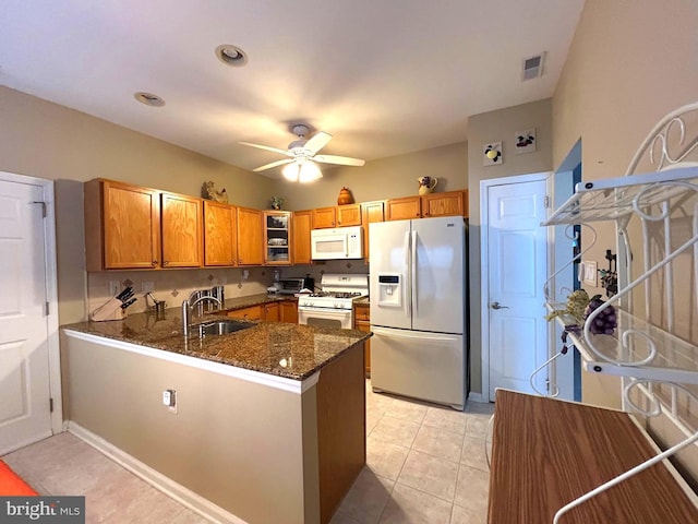 kitchen with a peninsula, white appliances, a sink, brown cabinetry, and dark stone countertops