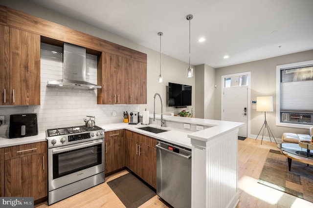 kitchen with a peninsula, a sink, appliances with stainless steel finishes, wall chimney range hood, and backsplash