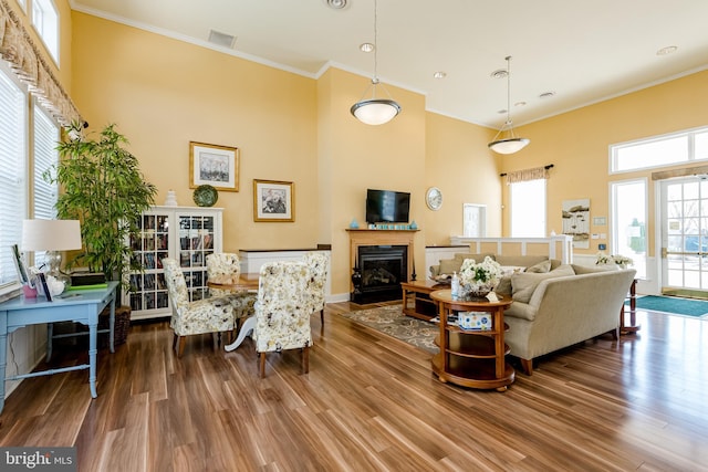 living area with visible vents, a towering ceiling, a fireplace with flush hearth, ornamental molding, and wood finished floors