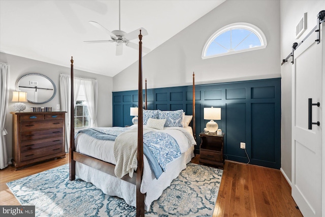 bedroom with a barn door, a ceiling fan, wood finished floors, high vaulted ceiling, and a decorative wall