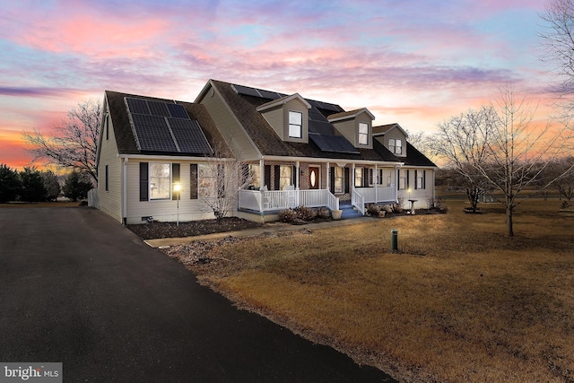 new england style home with driveway, a porch, roof with shingles, roof mounted solar panels, and a front lawn