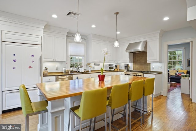 kitchen featuring wooden counters, dishwasher, white cabinetry, paneled refrigerator, and wall chimney exhaust hood