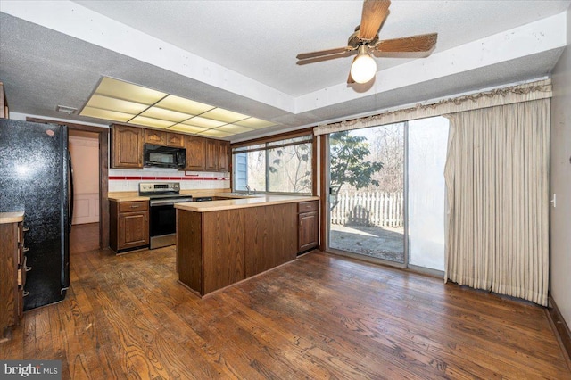 kitchen with a peninsula, a tray ceiling, black appliances, dark wood-type flooring, and light countertops