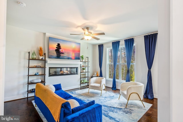 sitting room with a ceiling fan, a glass covered fireplace, dark wood-style flooring, and baseboards