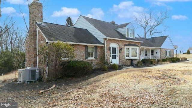 cape cod house with stone siding, a chimney, central AC, and roof with shingles