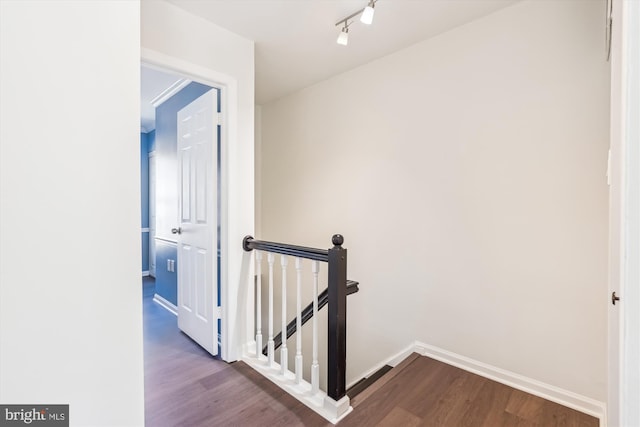 corridor featuring dark wood-type flooring, rail lighting, baseboards, and an upstairs landing