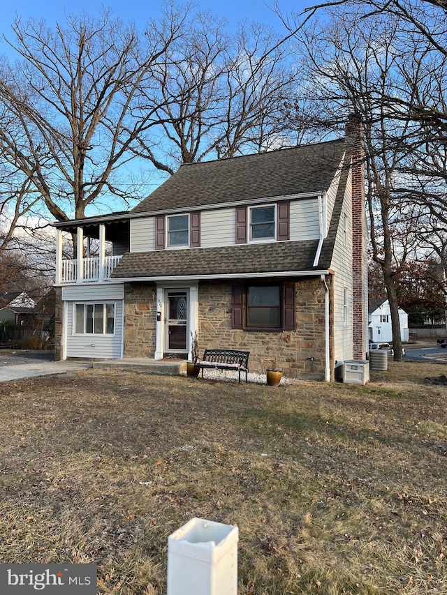 view of front facade with stone siding, roof with shingles, a chimney, and a balcony