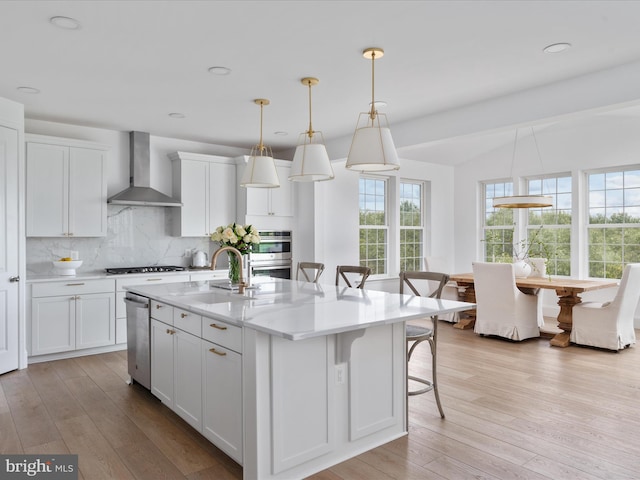 kitchen featuring a breakfast bar, white cabinetry, hanging light fixtures, a kitchen island with sink, and wall chimney range hood