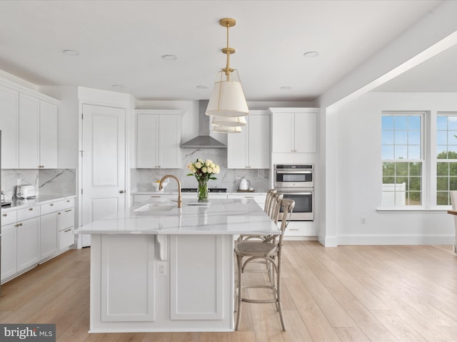 kitchen featuring stainless steel double oven, decorative light fixtures, an island with sink, and wall chimney exhaust hood