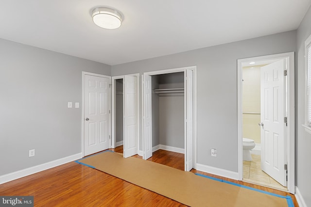 unfurnished bedroom with ensuite bath, wood finished floors, two closets, and baseboards